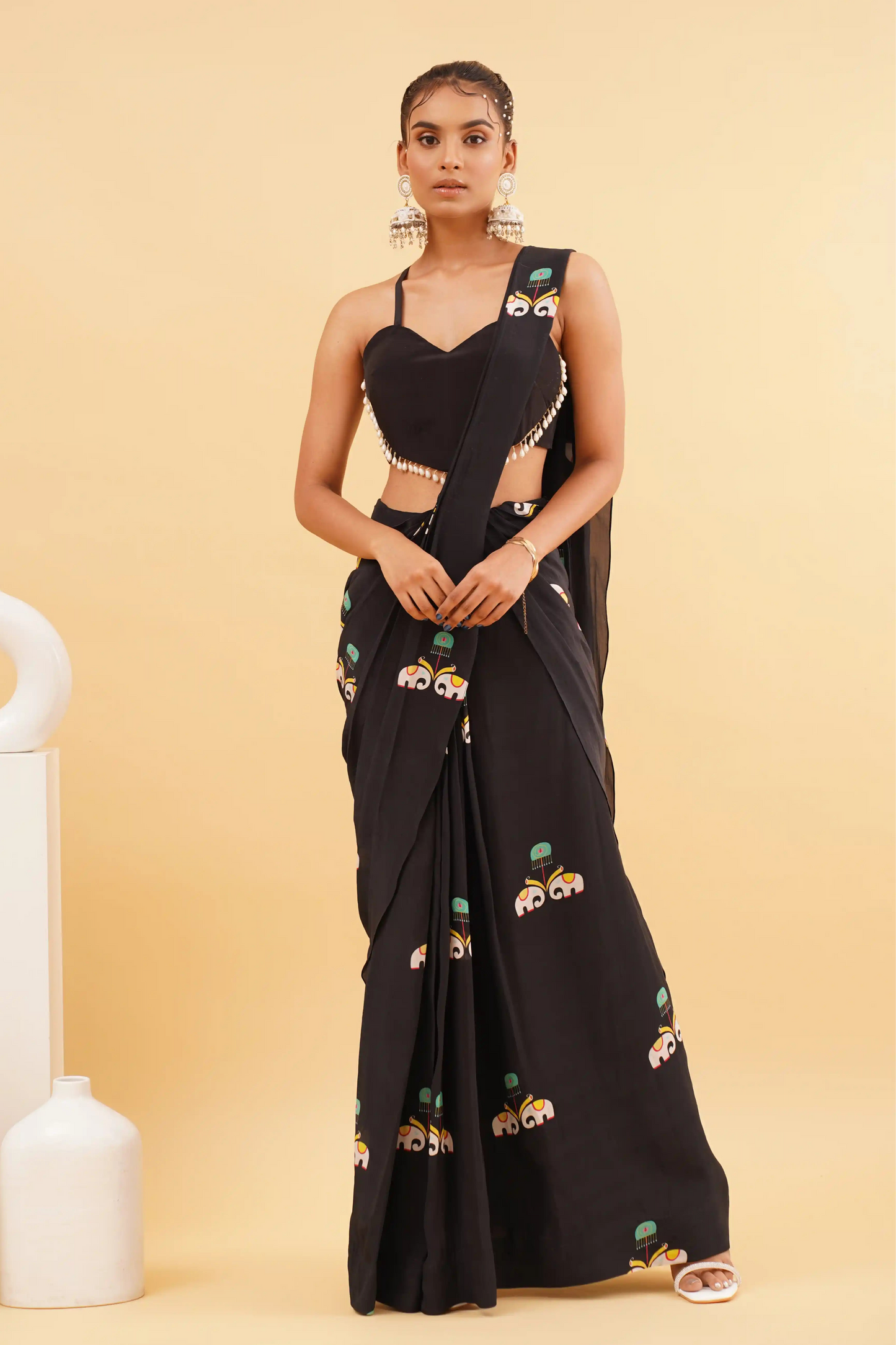 A woman standing in front of a black saree with a floral print on it, posing against a yellow background with white decorative items in the background.