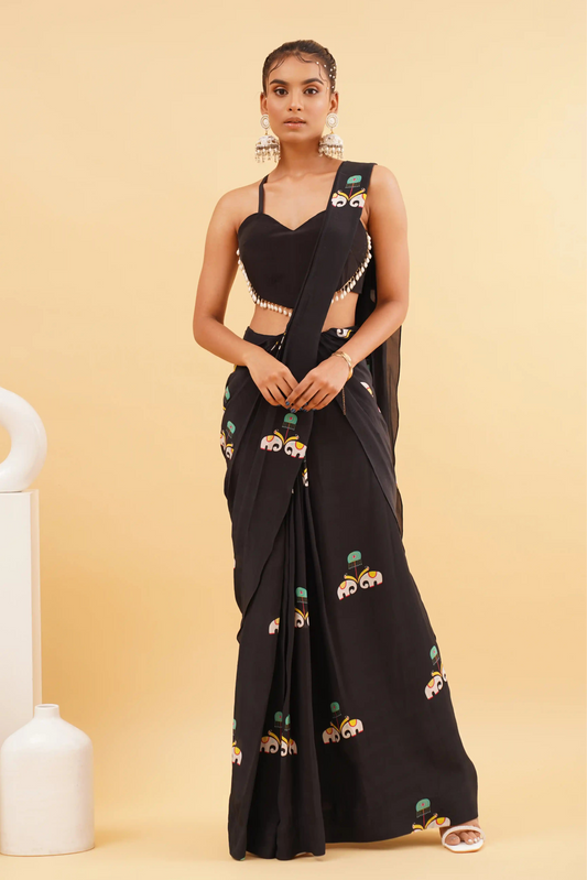 A woman standing in front of a black saree with a floral print on it, posing against a yellow background with white decorative items in the background.