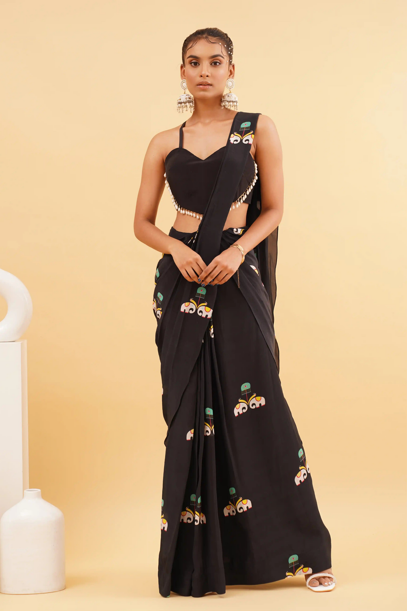 A woman standing in front of a black saree with a floral print on it, posing against a yellow background with white decorative items in the background.