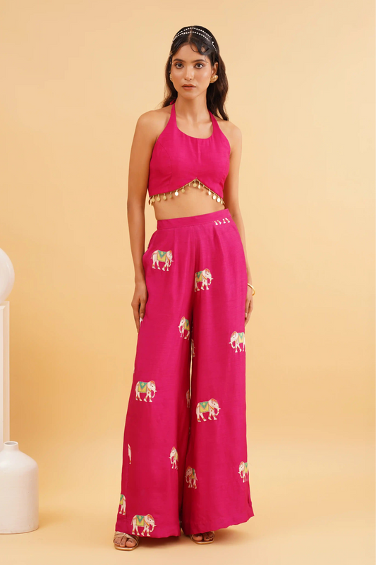 A woman wearing a vibrant fuchsia pink Nazrana co-ord set with golden elephant motifs, featuring a structured silhouette and tailored fit, standing next to white column-like decorative objects.