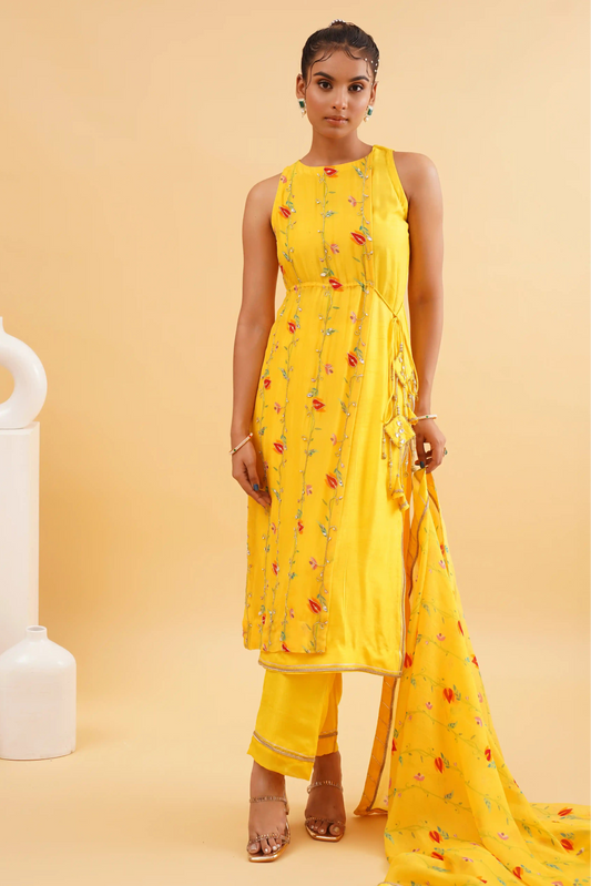 A woman wearing a vibrant yellow sleeveless kurta set with floral embroidery and organza dupatta, standing against a yellow background with white decor.