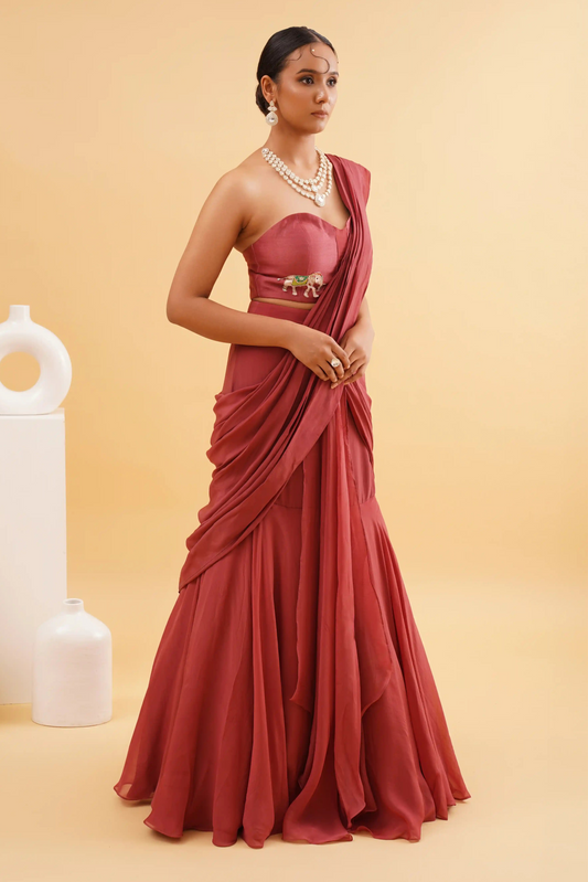 A woman modeling a rust-colored Anmol saree with intricate elephant embroidery, accompanied by a red and gold blouse piece, with a modern presentation in a studio setting.