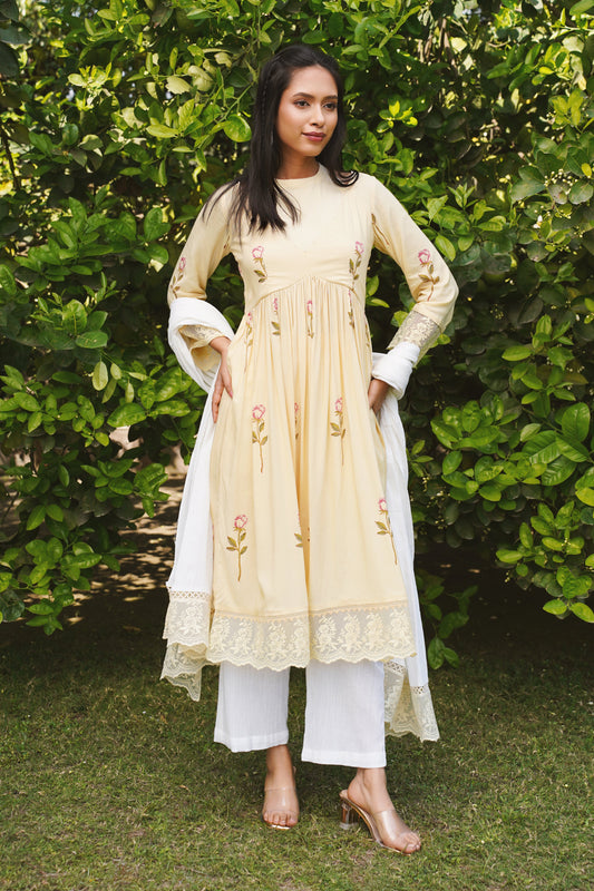 A woman standing outdoors wearing a floral kurta set with lace and pearl detailing on the neckline, paired with a flowy dupatta and white pants.