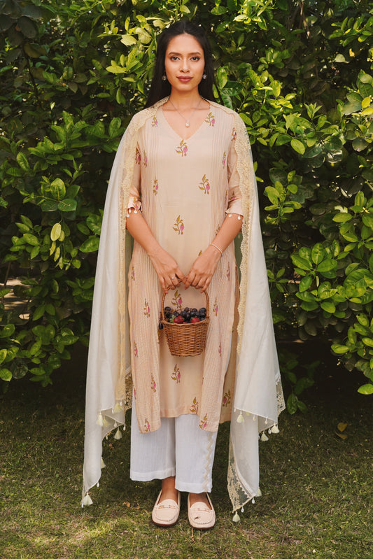 A woman wearing a beige kurta set with rosebud print, pin-tuck pleats, and lace accents, paired with a dupatta. She is standing against a lush green background.