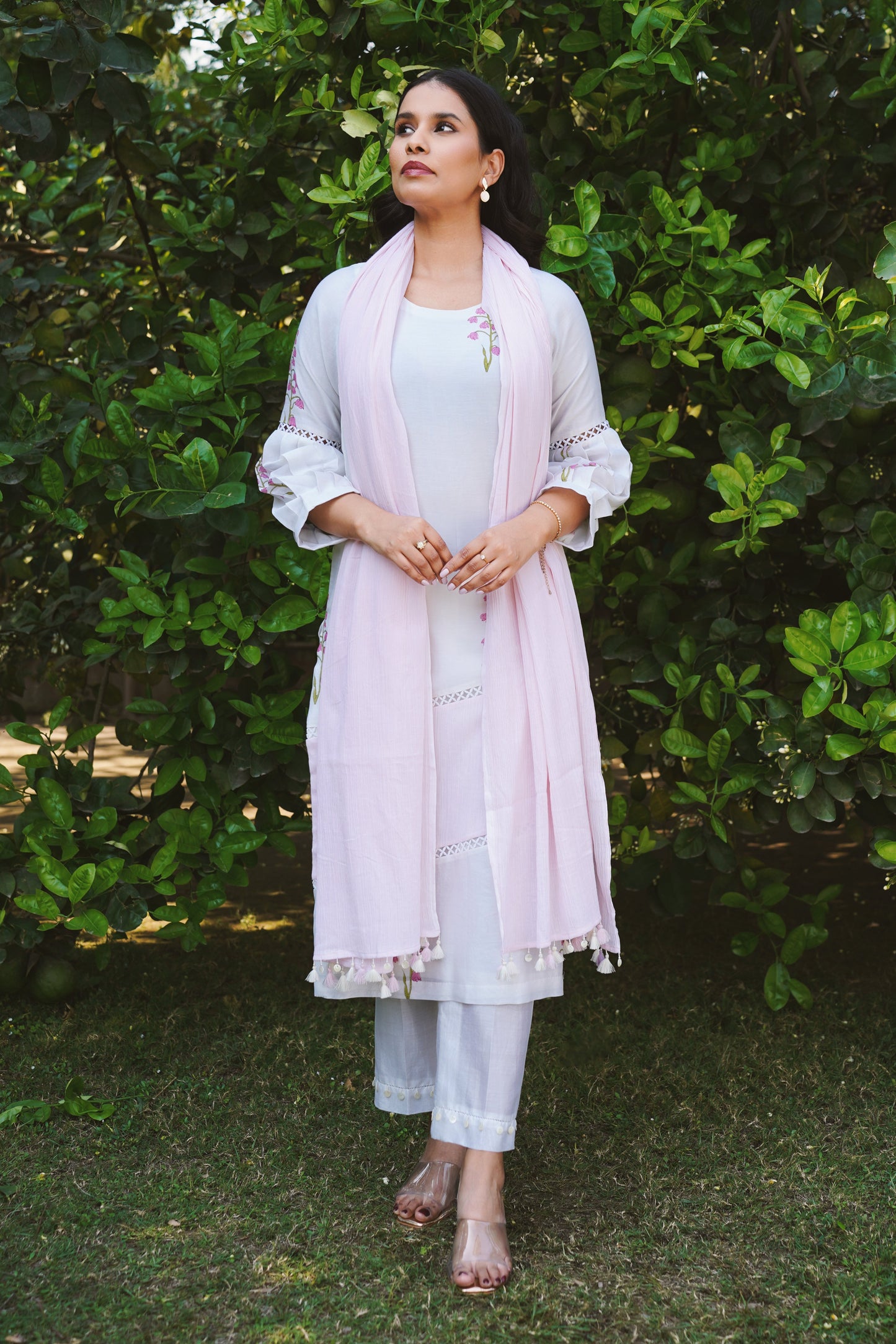 A woman wearing a white and pink kurta set with cherry blossom print, tailored pants, and a dupatta standing against a lush green background.