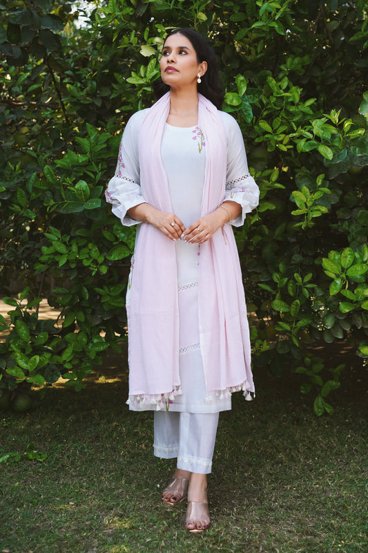 A woman wearing a white and pink kurta set with cherry blossom print, tailored pants, and a dupatta standing against a lush green background.