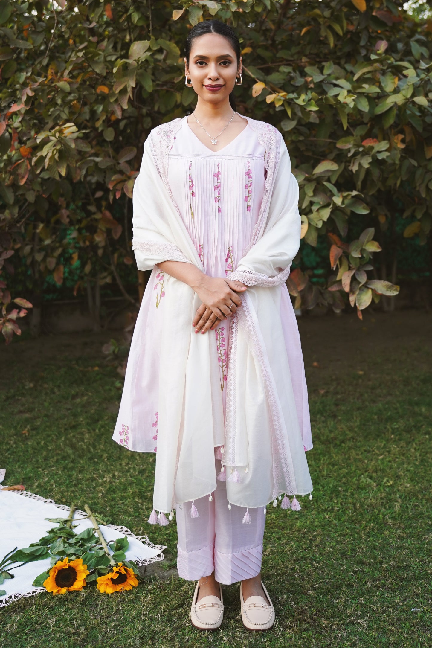 A woman standing outdoors wearing a blush pink kurta set with pleated details at the yoke and pin-tucked pants, accompanied by a lace-trimmed dupatta.
