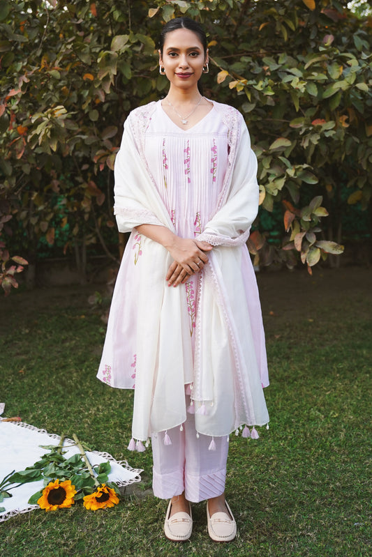 A woman standing outdoors wearing a blush pink kurta set with pleated details at the yoke and pin-tucked pants, accompanied by a lace-trimmed dupatta.