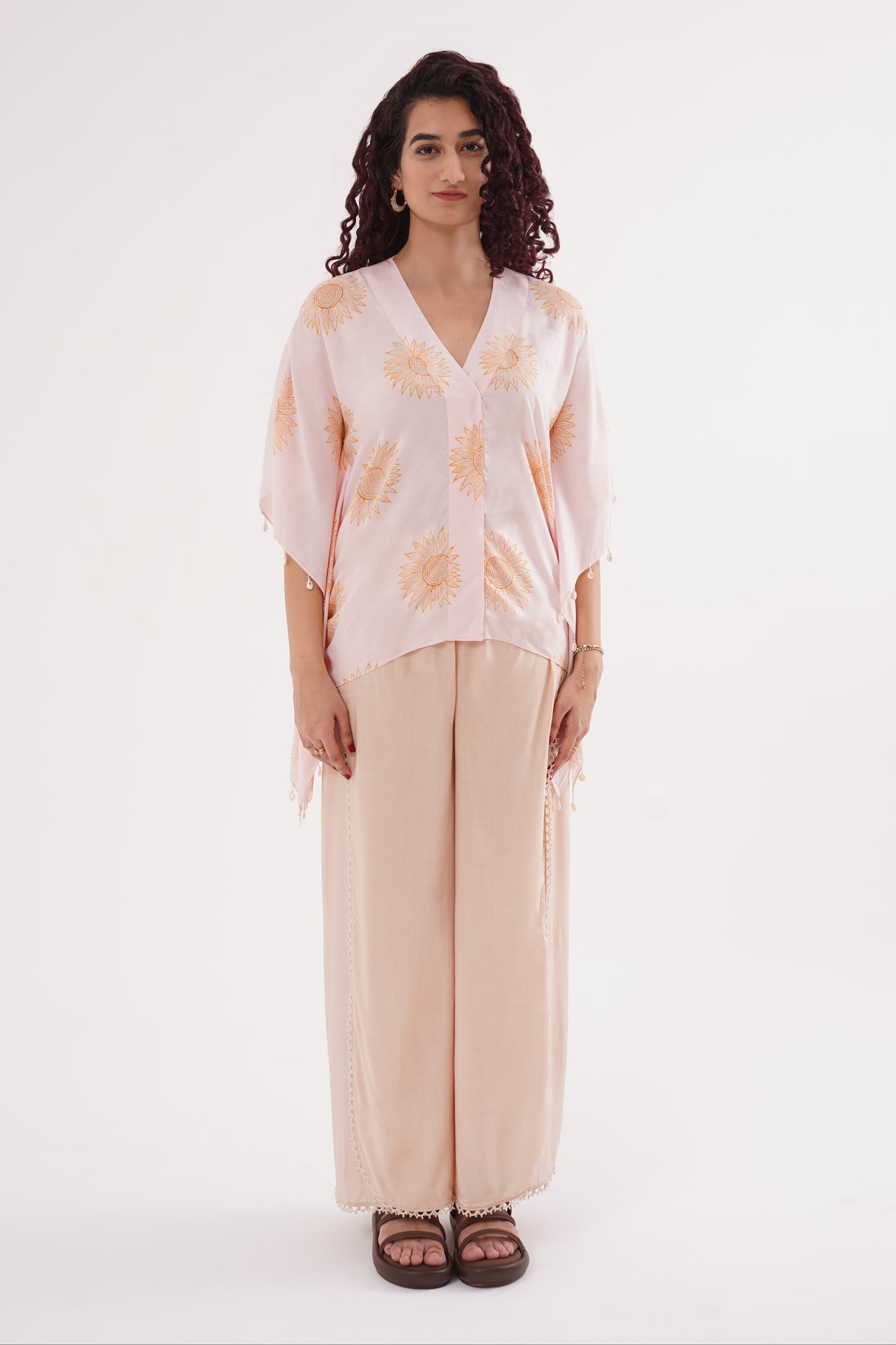 A female model wearing a sunflower print kaftan set with shell-button details on the sleeves and wide-leg pants with flap accents. The set is in blush pink and soft caramel colors.