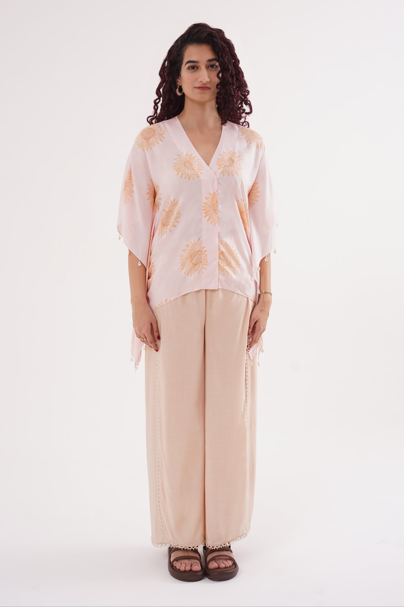 A female model wearing a sunflower print kaftan set with shell-button details on the sleeves and wide-leg pants with flap accents. The set is in blush pink and soft caramel colors.